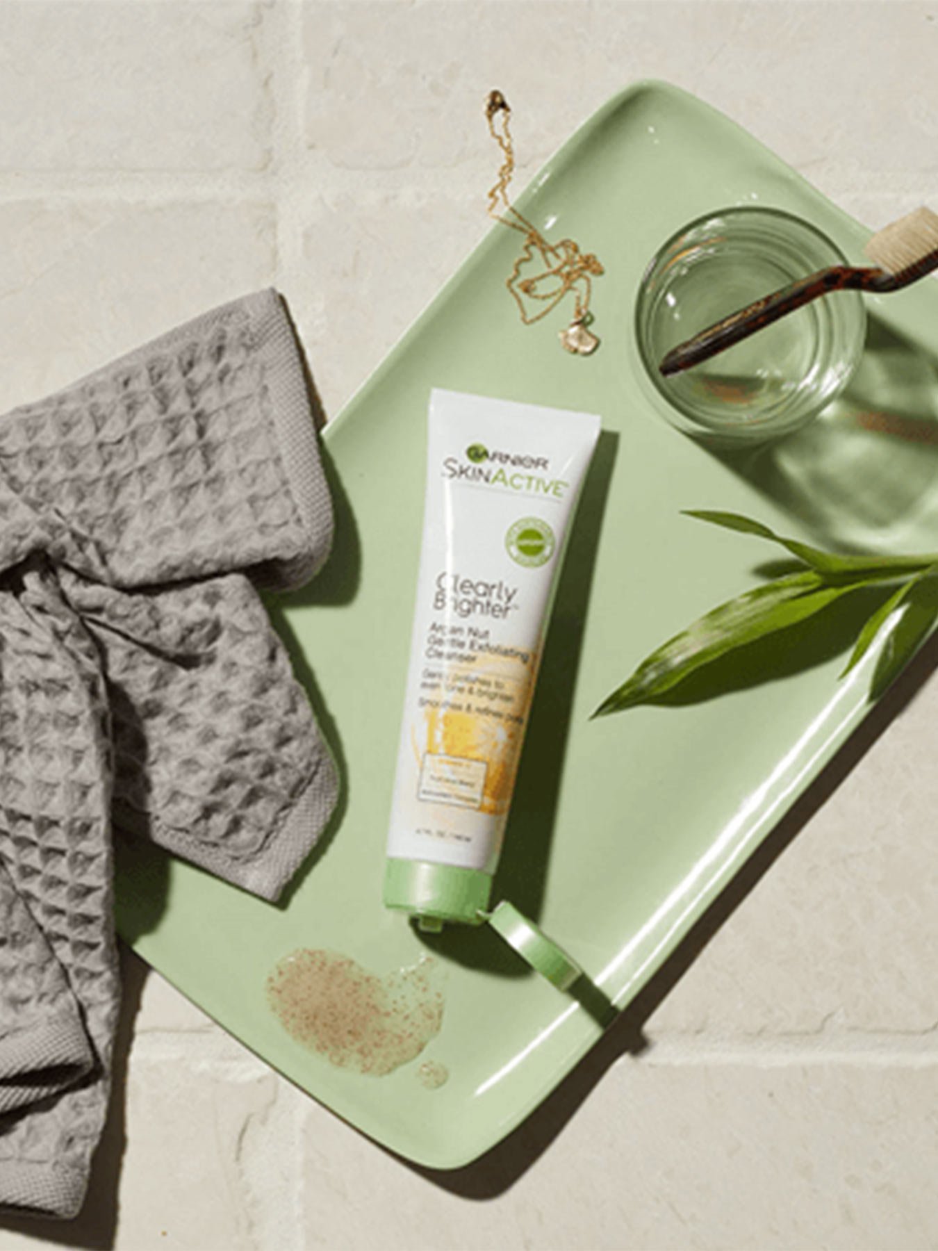 Clearly Brighter Argan Nut Gentle Exfoliating Cleanser open with some product spilled on a green tray holding a glass with a toothbrush, a gold necklace and pendant, a leaf, and a grey washcloth.
